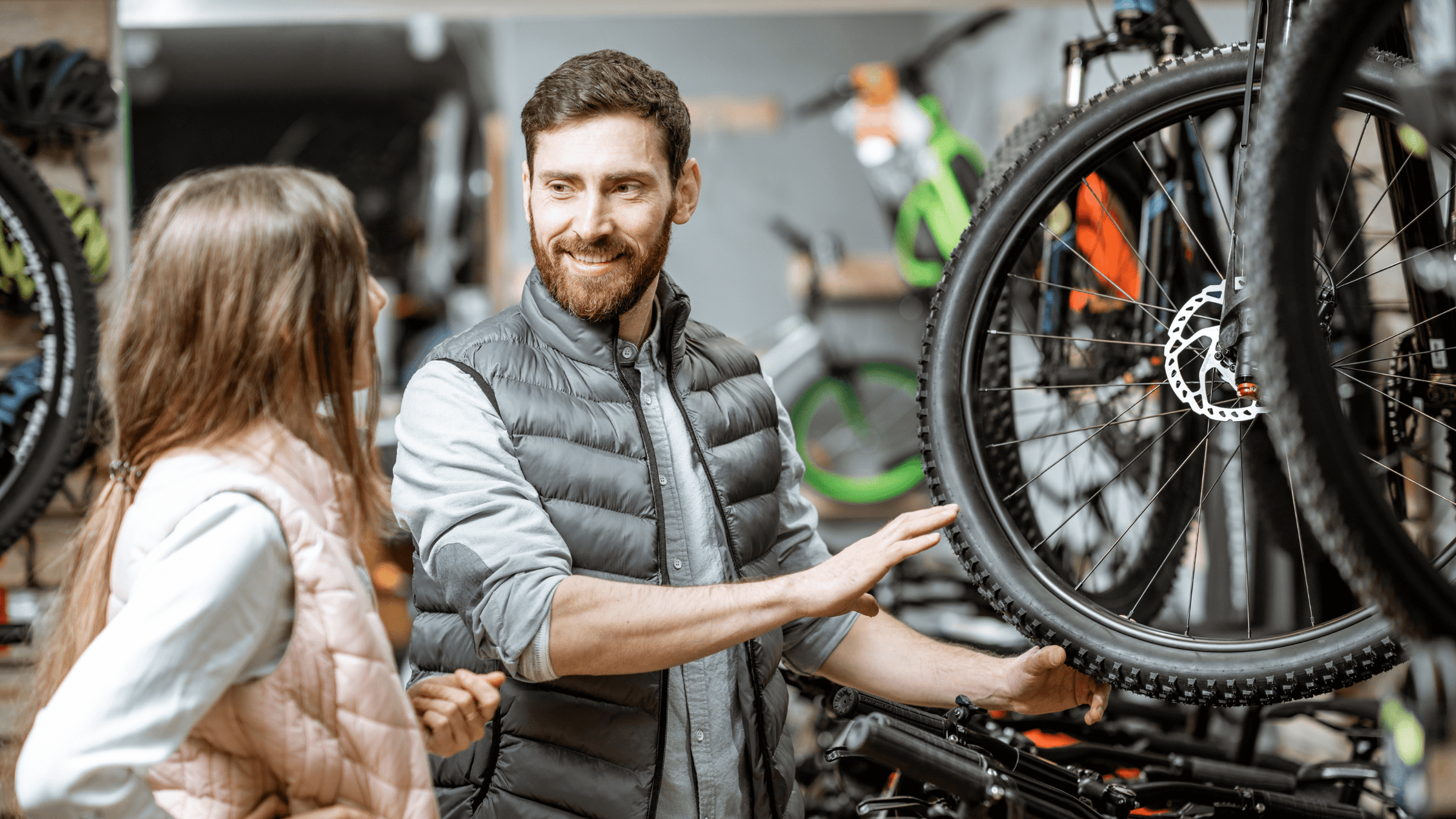Ein Verkäufer zeigt einer Kundin ein Mountainbike in einem Fahrradgeschäft. Beide unterhalten sich freundlich, während der Verkäufer das Vorderrad des Fahrrads erklärt. Im Hintergrund sind weitere Fahrräder und Zubehör zu sehen.